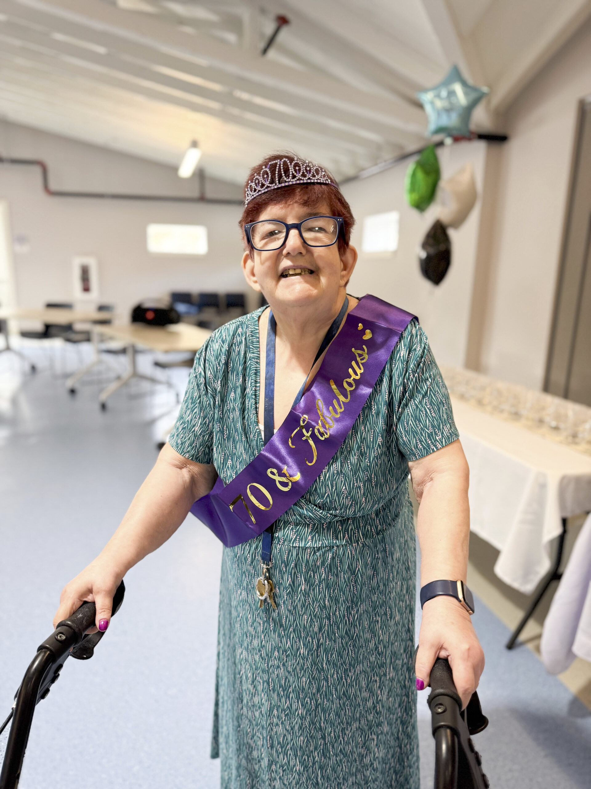 Woman standing holding a walker, wearing a sash and birthday crown.
