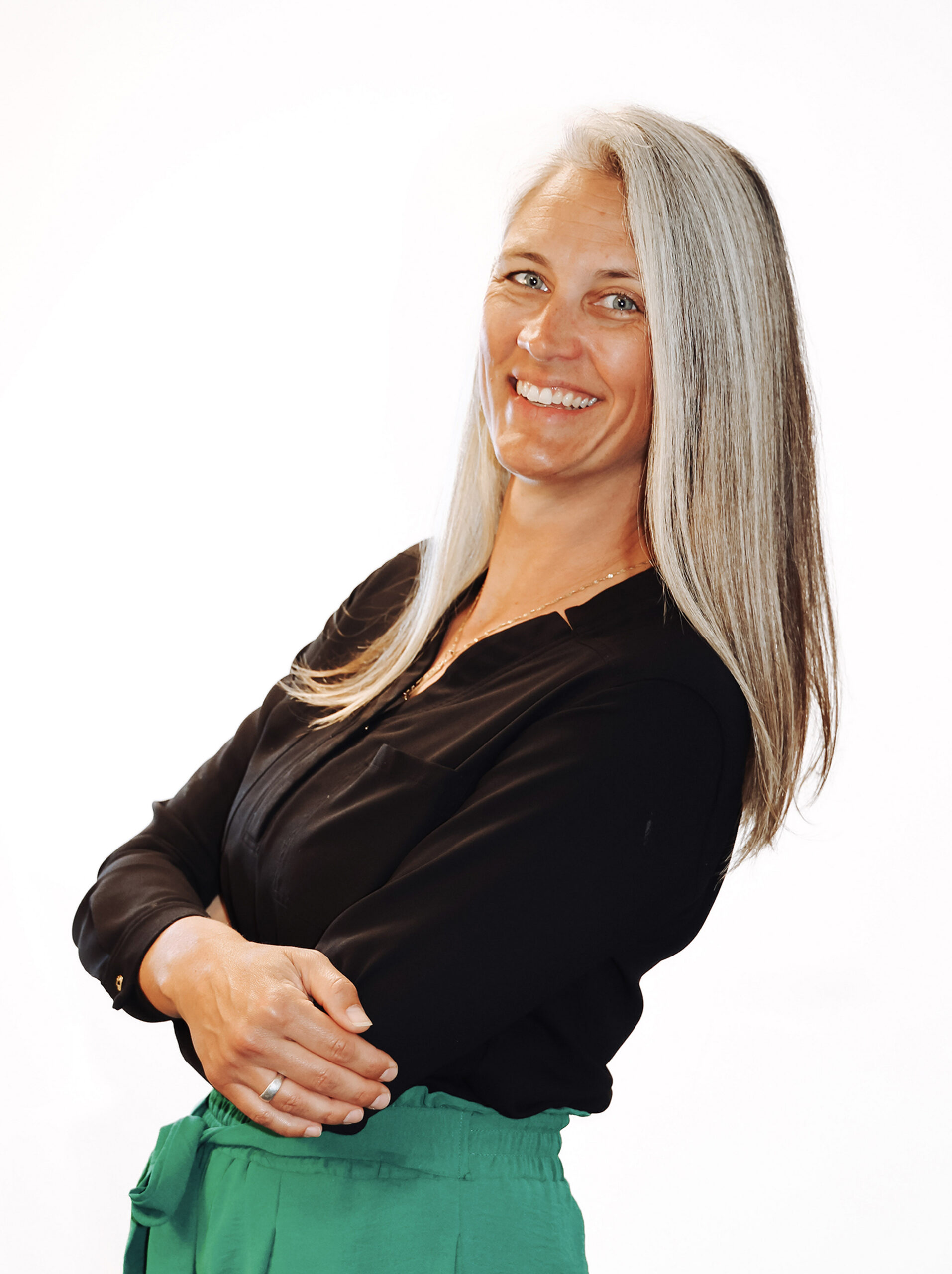 Woman standing at a white backdrop wearing a black top and green pants.